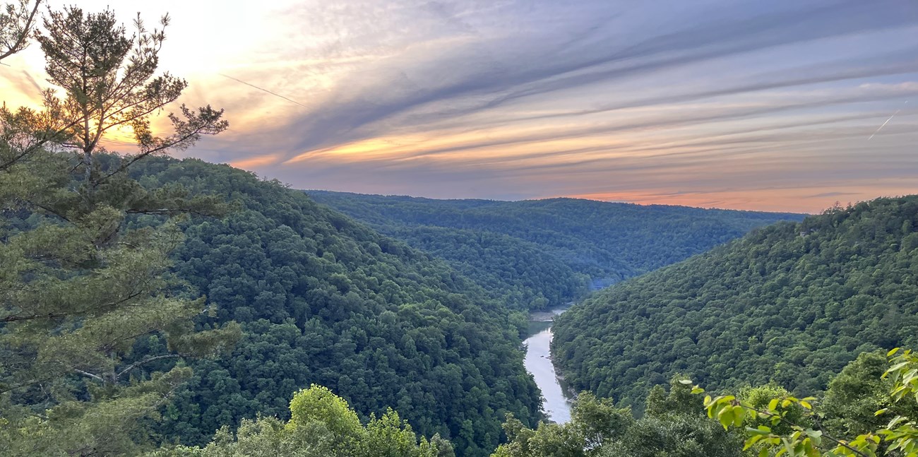 An aerial view of verdant, forested mountains bisected by river, with the sun setting behind swirling, orange-ish clouds in the distance.