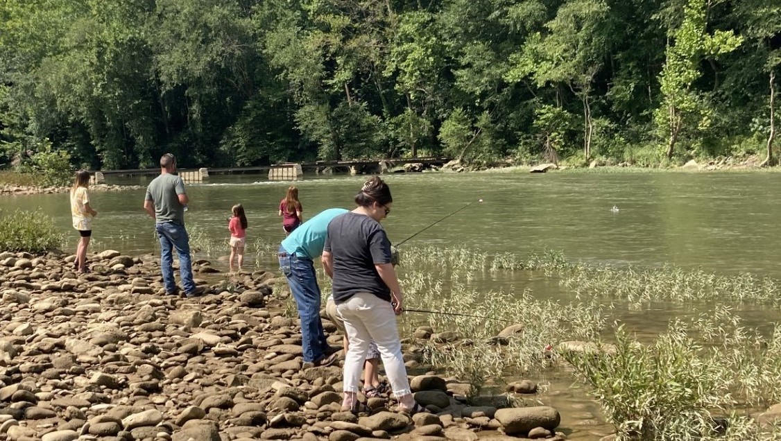 Children and adults standing on a pebble-strewn riverbank with the green river itself and the other forested bank beyond them.