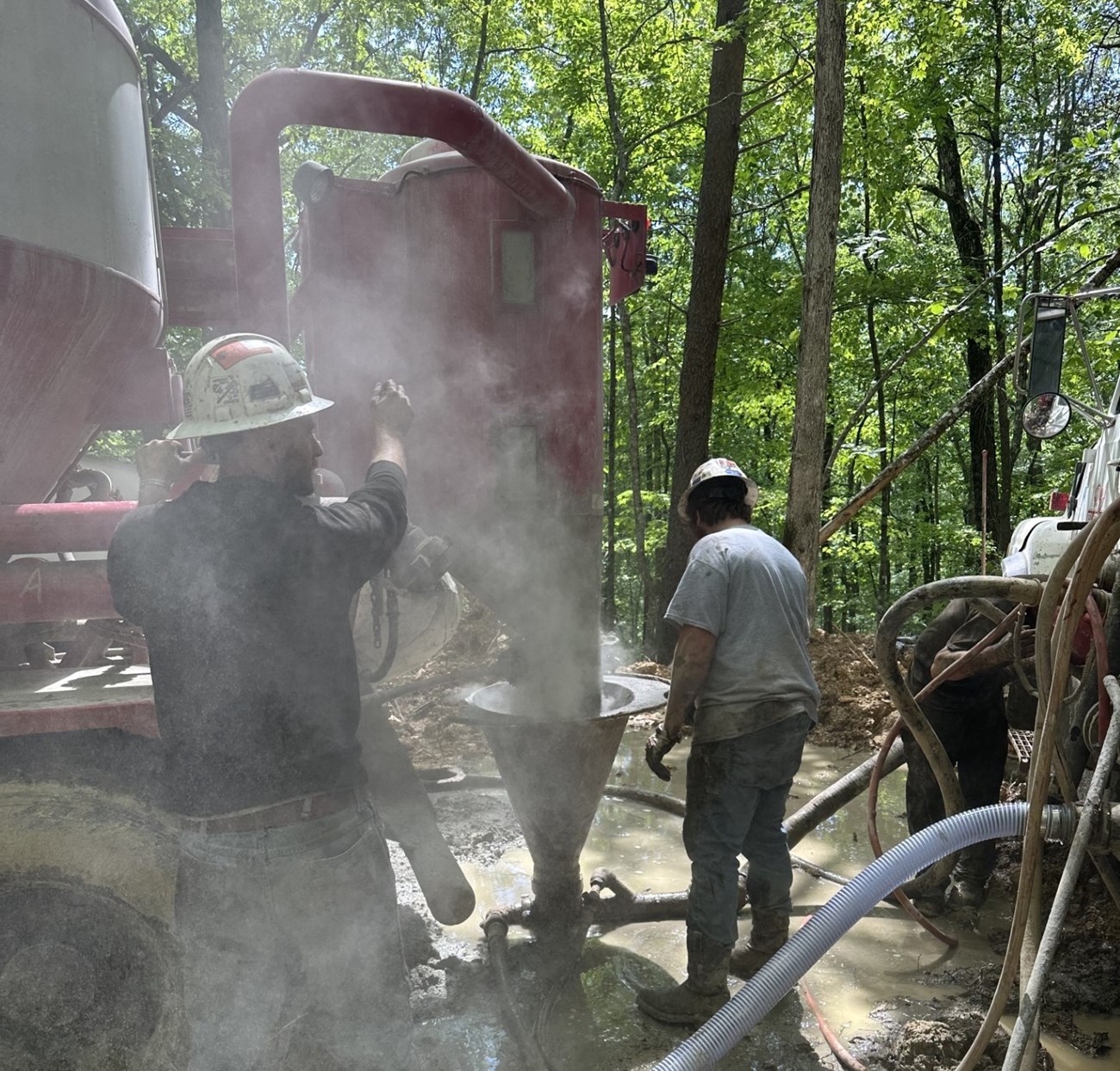 Two men wearing work helmets funnel cement from a truck into an abandoned oil well pipe with all sorts of related tubing nearby and a stand of trees in the background.