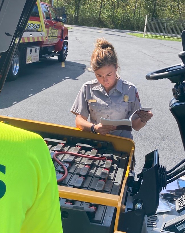 Female uniformed ranger stands looking down at a book, in front of the open hood of a yellow and black forklift.