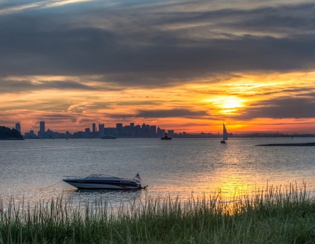 A sunset fills a cloudy sky with city buildings and sailboat on the horizon. There is calm water, a moored boat, and grasses in the foreground.