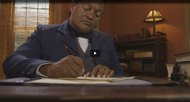 Screenshot of a video with a living historian portraying Booker T. Washington writing at a desk