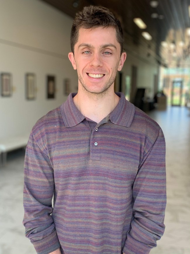 White man with short brown hair and stubble wearing a purple shirt.