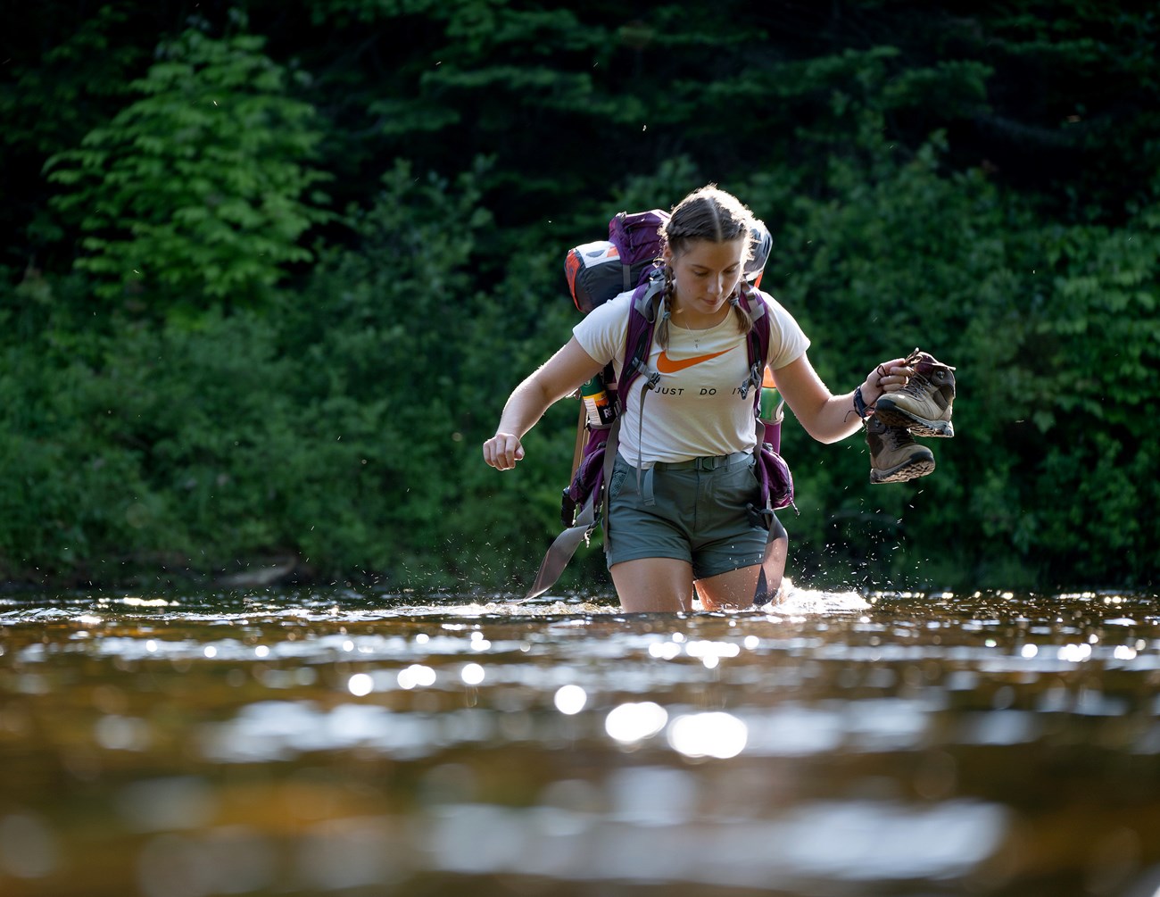 A hiker with a backpack holds their hiking boots above the water. The water is thigh high.