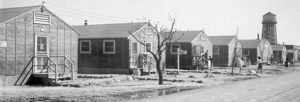 Black and white image of a Minidoka residential block, with a few people standing outside.