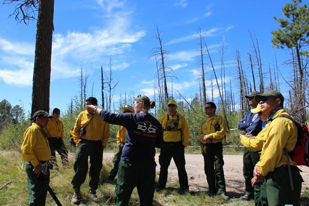 A group of people outdoors in yellow shirts gathered around a woman who is pointing at something in the distance.