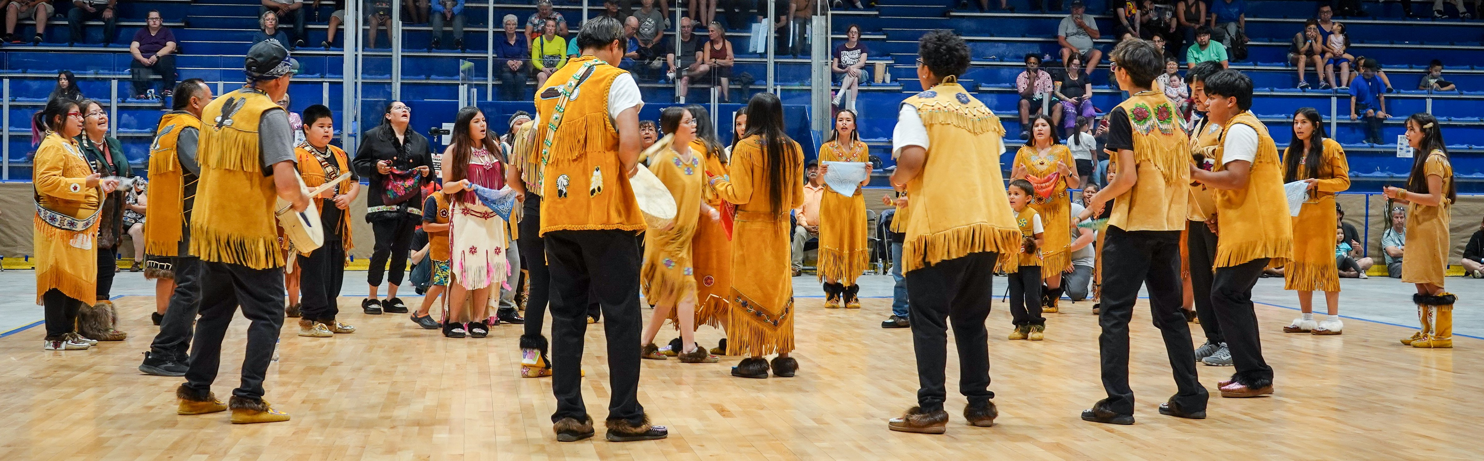 a group of Alaska Native performers wearing traditional clothing gather on a dance floor.
