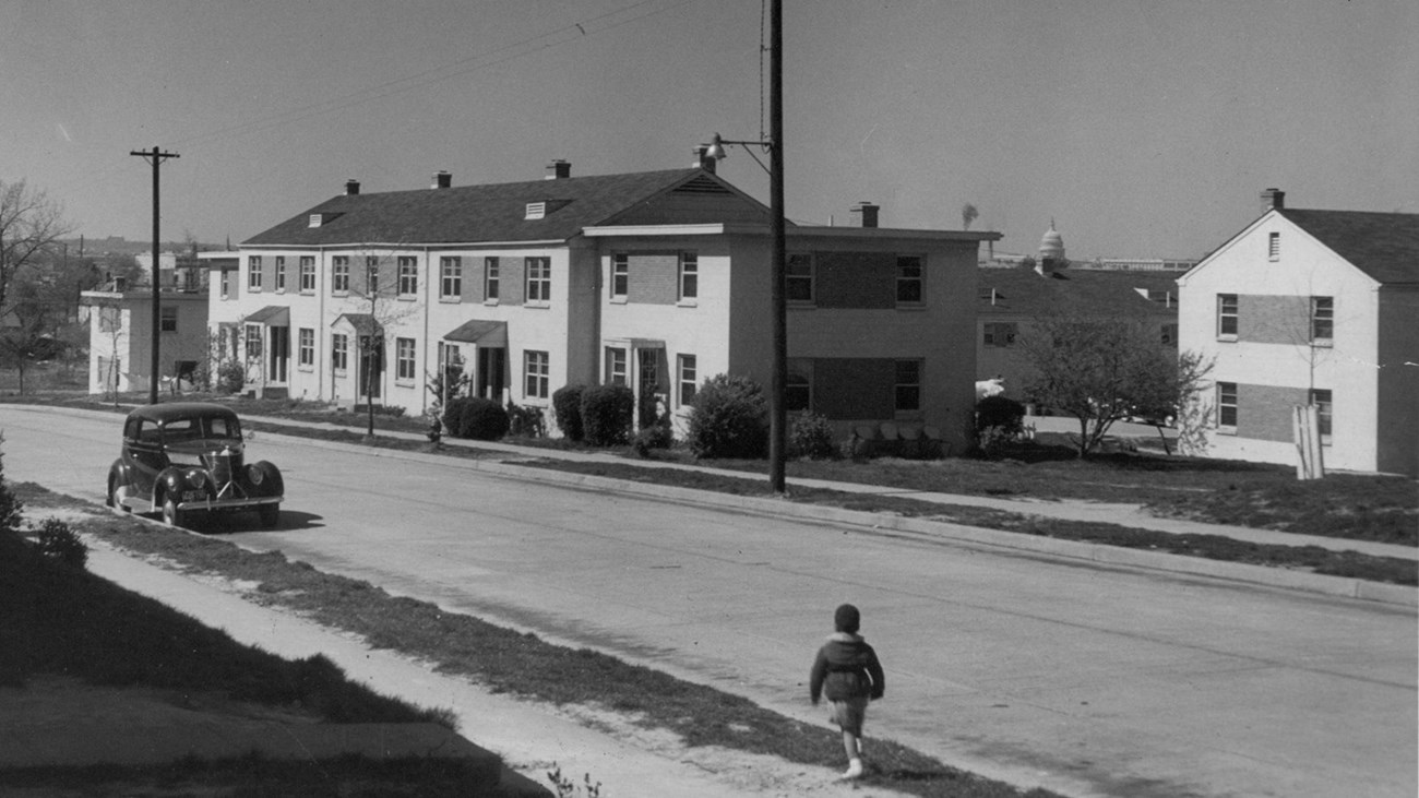 1940s photo of street with 2 story white buildings, parked car, and child walking next to street.