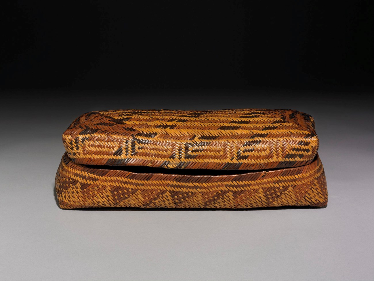 An intricate woven lidded basket sits against a gray background. 