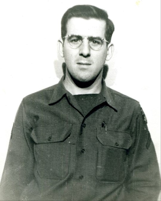 An image of a man in uniform standing against a wall. He has short brown hair and is wearing glasses.