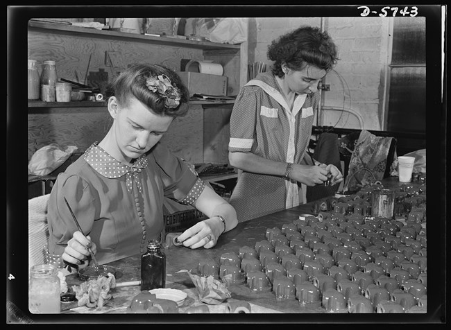 Two young women paint Y-shaped metal parts at a table with finished parts arranged in front of them. At left, Belva is seated & wears a top with polka dots & a flower clip in her hair. At right, Henriette is standing & has dark curly hair & wears a dress