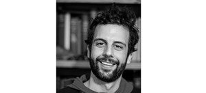 A dark-haired young man smiling at the camera in front of a bookcase of books