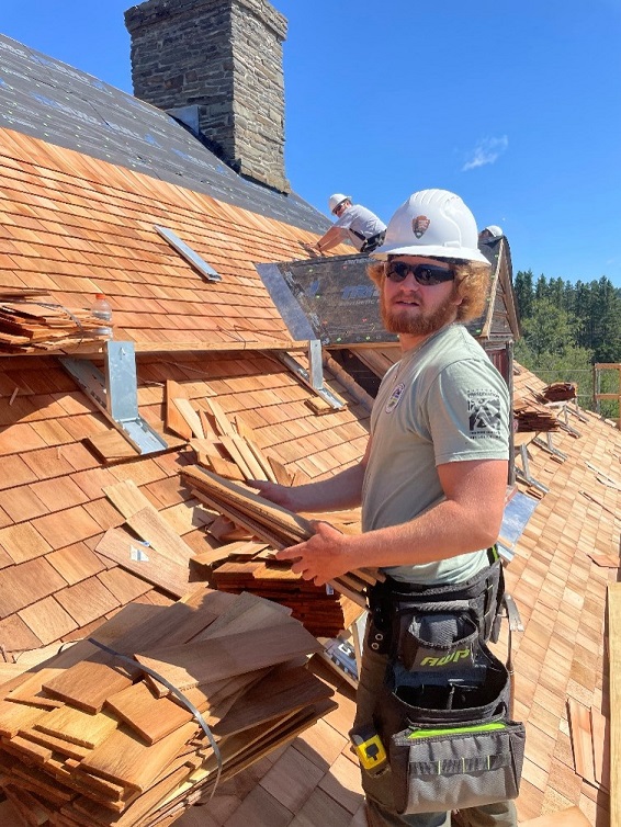 Employee wearing sunglasses and white hardhat holds roofting tiles while working atop a building.