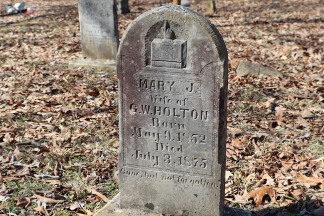 A gray tombstone features a symbol of a Bible and a hand pointing up.