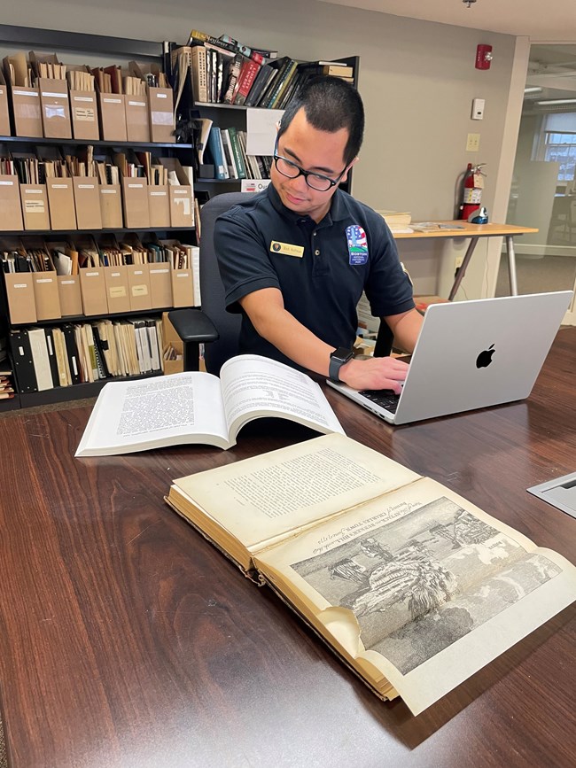 A young man referencing a book while typing on a laptop.