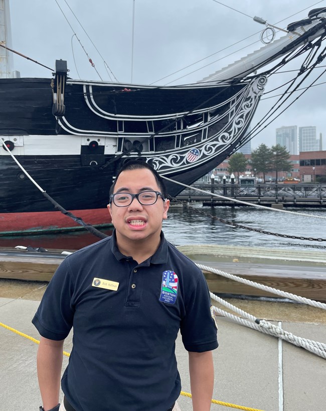 A young man smiling in front of the USS Constitution ship at Boston National Historical Park.