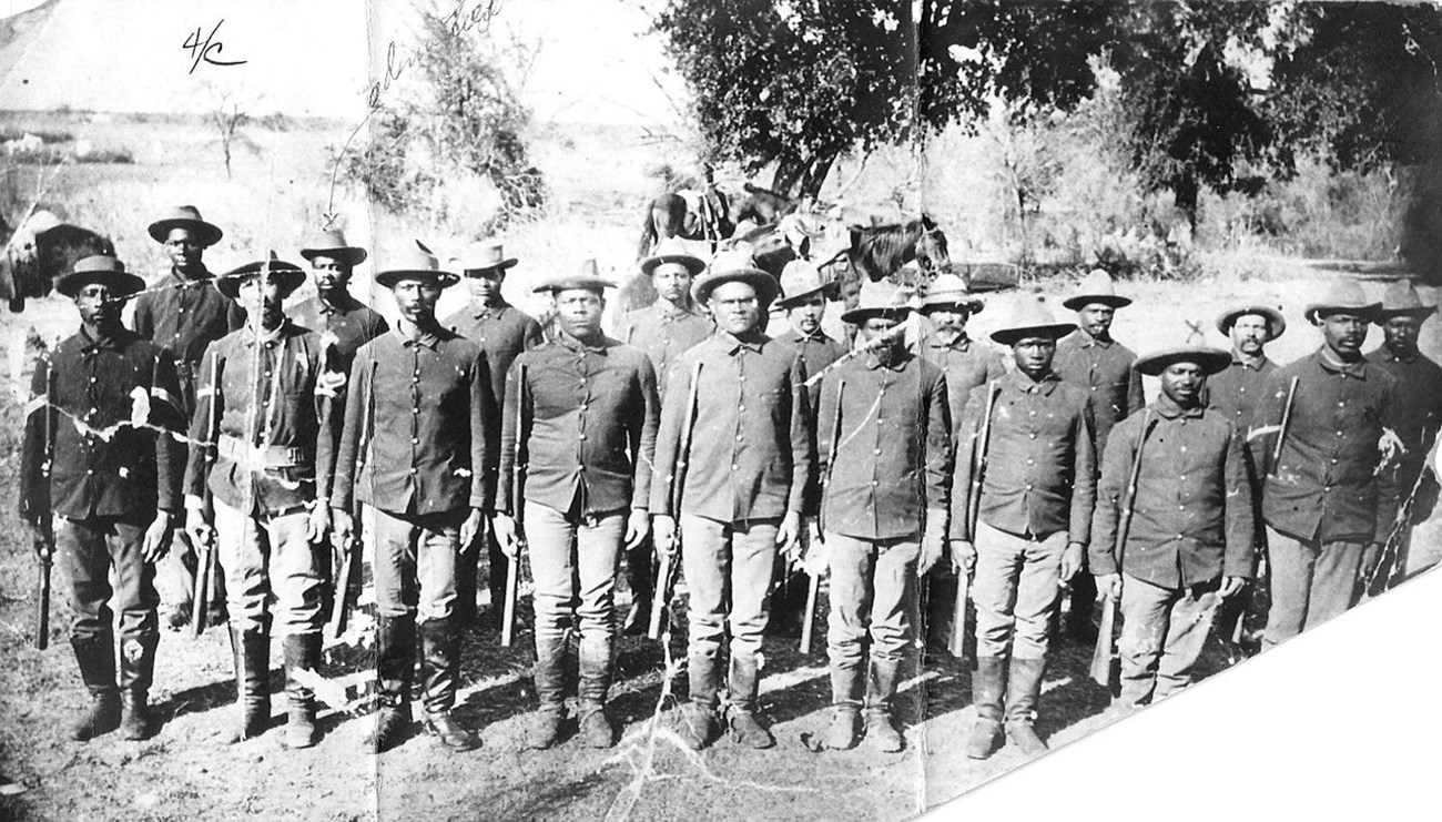 Black and white phots of two rows of Black Seminoles standing at attention and looking directly at the viewer. They are all wearing the US Army uniform of the 1890s with black riding boots that come up to their knee. They are holding their carbines.
