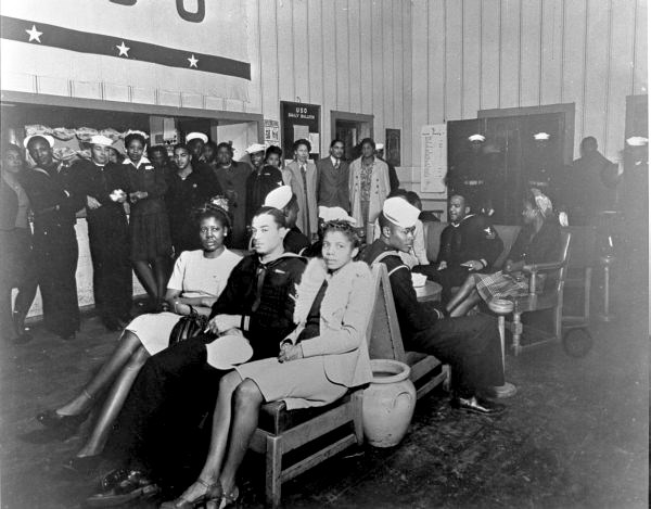 Black and white photo of an African American man in uniform sitting between two African American women. African American men and women stand around the perimeter of the room, also in uniform