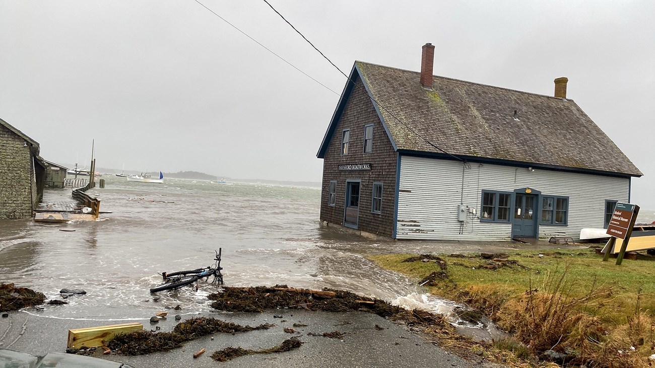 A small wooden building covered in shingles is affected by storm tides. The water completely encroaches on the base, and reaches up the road.