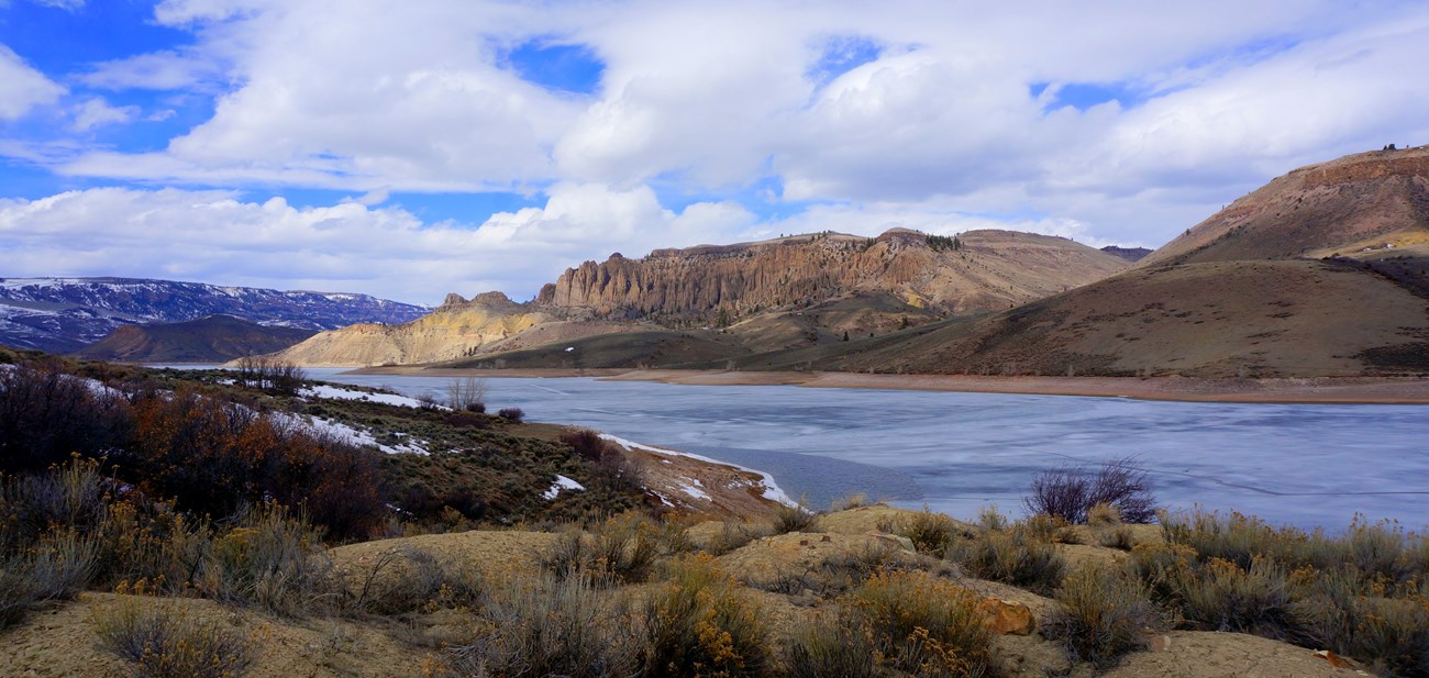 The Blue Mesa Reservoir in March, with ice cover remaining on the surface of the reservoir and some small snowpacks scattered along the shoreline and on distant mesas.