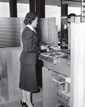A woman faces away from us towards a counter. She is wearing a dark colored uniform and pumps.