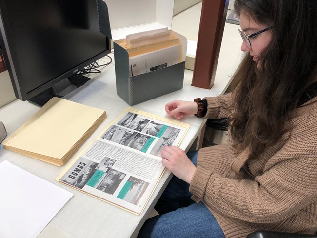 A woman with dark hair sits at a desk turning the pages of a "Bos'n's Whistle" magazine.
