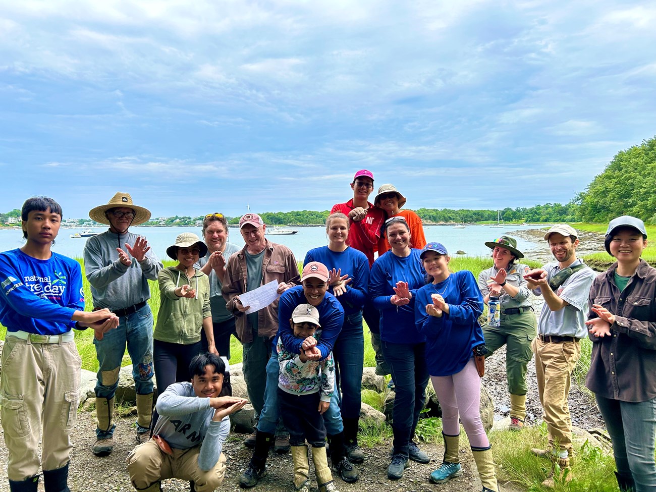 A group of 16 people smile at the camera. Each is putting their hands together to resemble clamshells. There is open water and forested shoreline behind them.