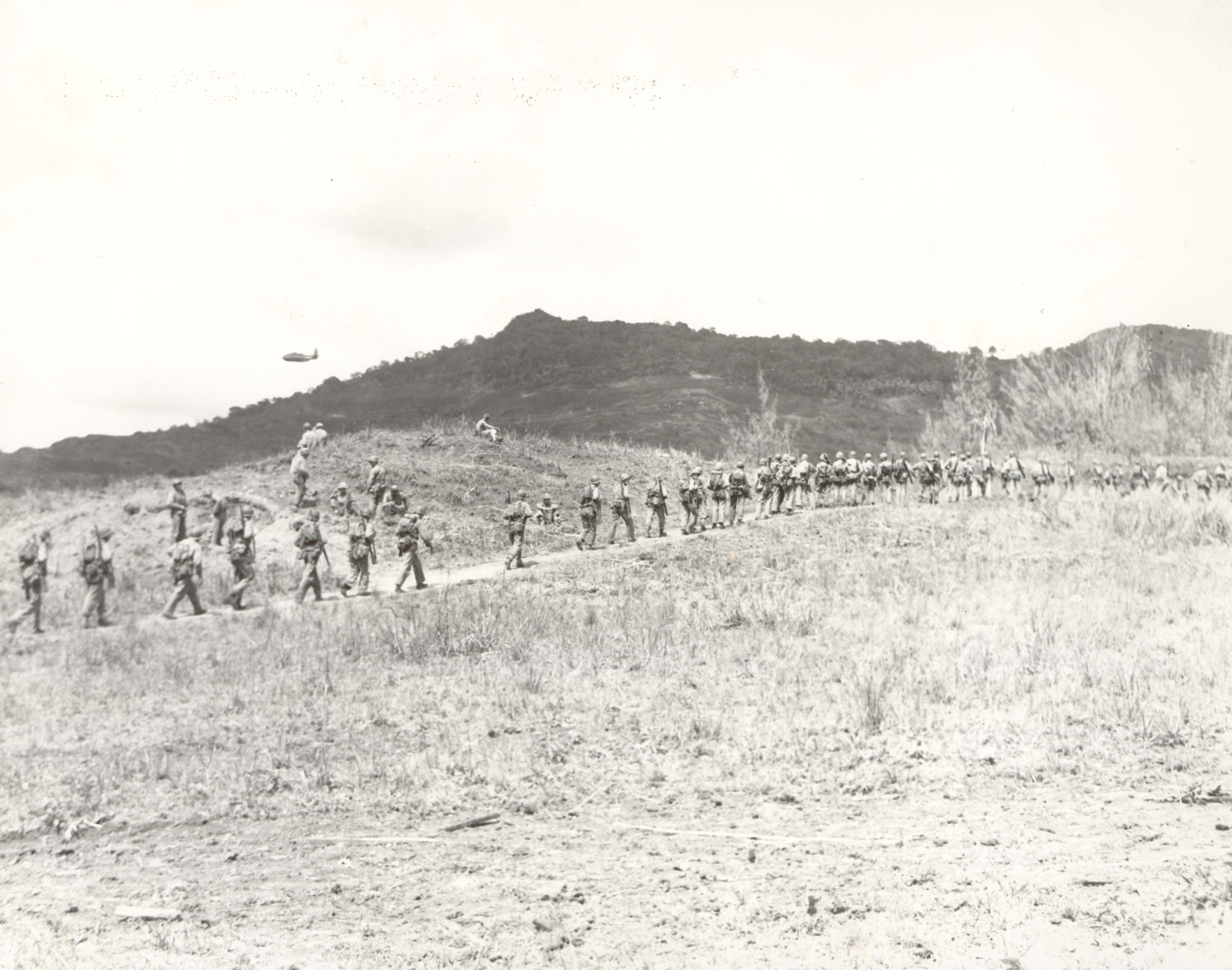 Soldiers march in a single file across a barren landscape, with rugged hills in the background. A plane flies low in the background.