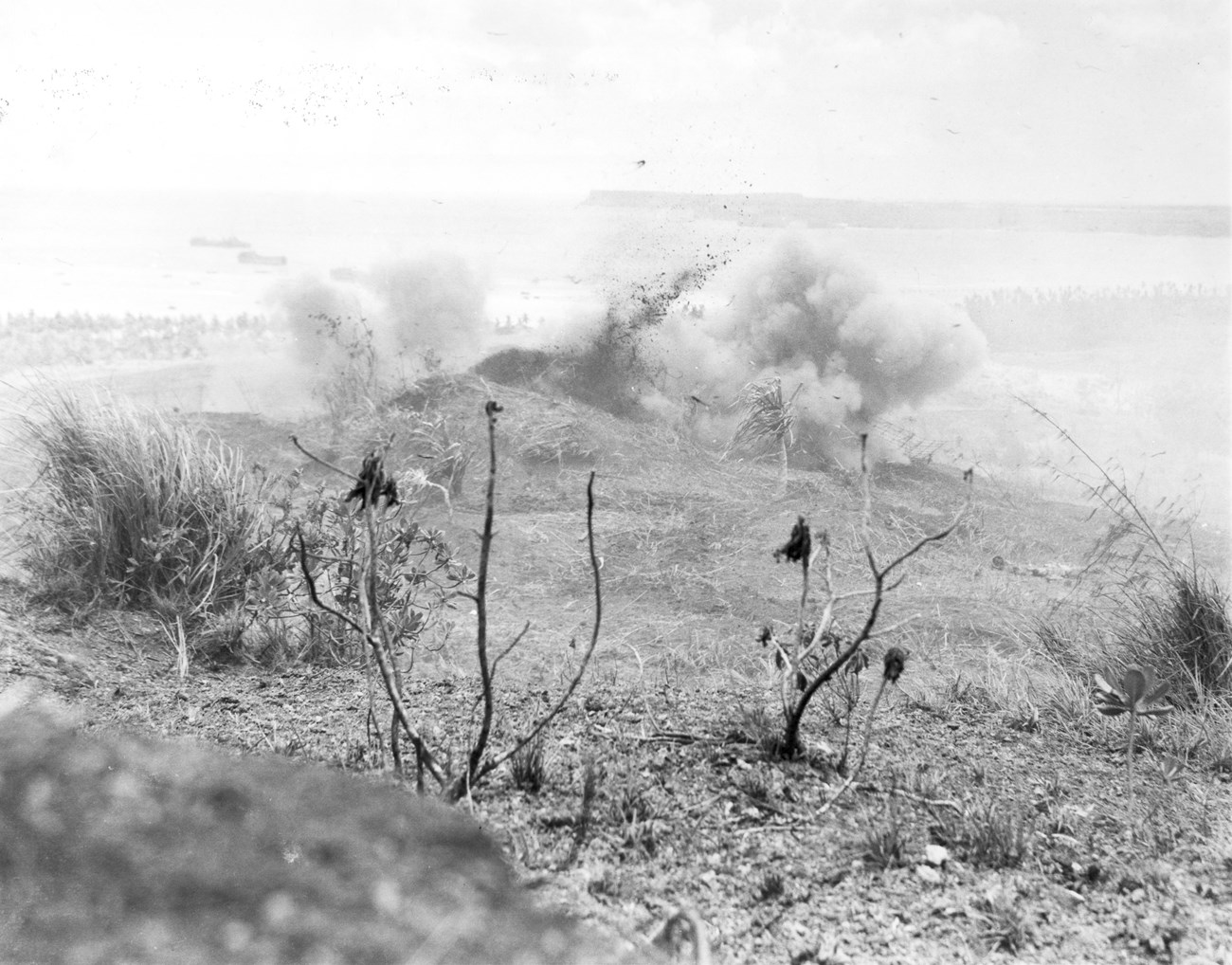 A barren, battle-scarred hill with an explosion creating a cloud of smoke. Sparse vegetation in the foreground.