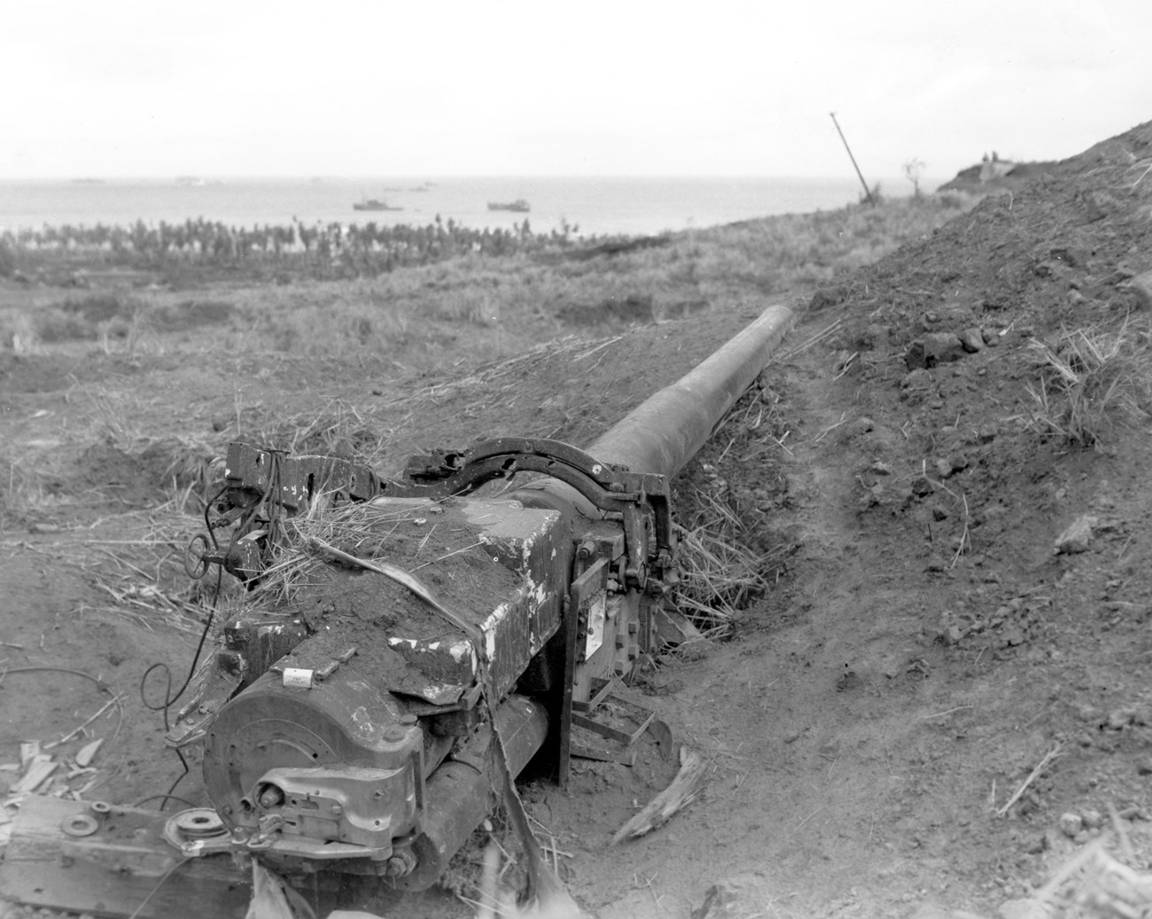A rusted artillery piece lies in a muddy trench, overlooking a tranquil sea with distant boats and a crowd of soldiers in the background.