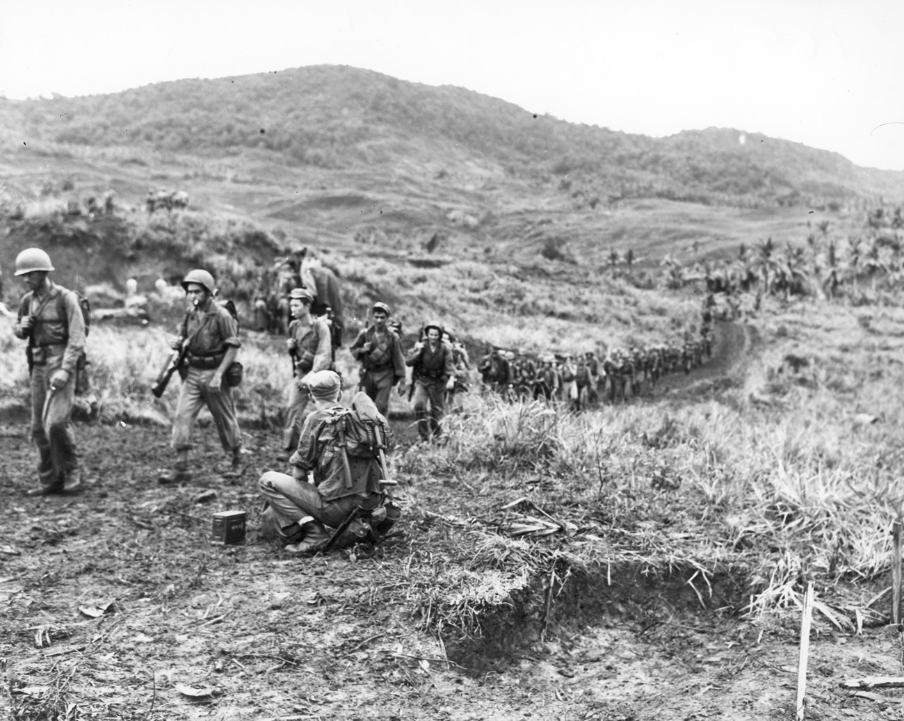A long line of soldiers in helmets marches through a muddy, grassy landscape with rolling hills in the background.