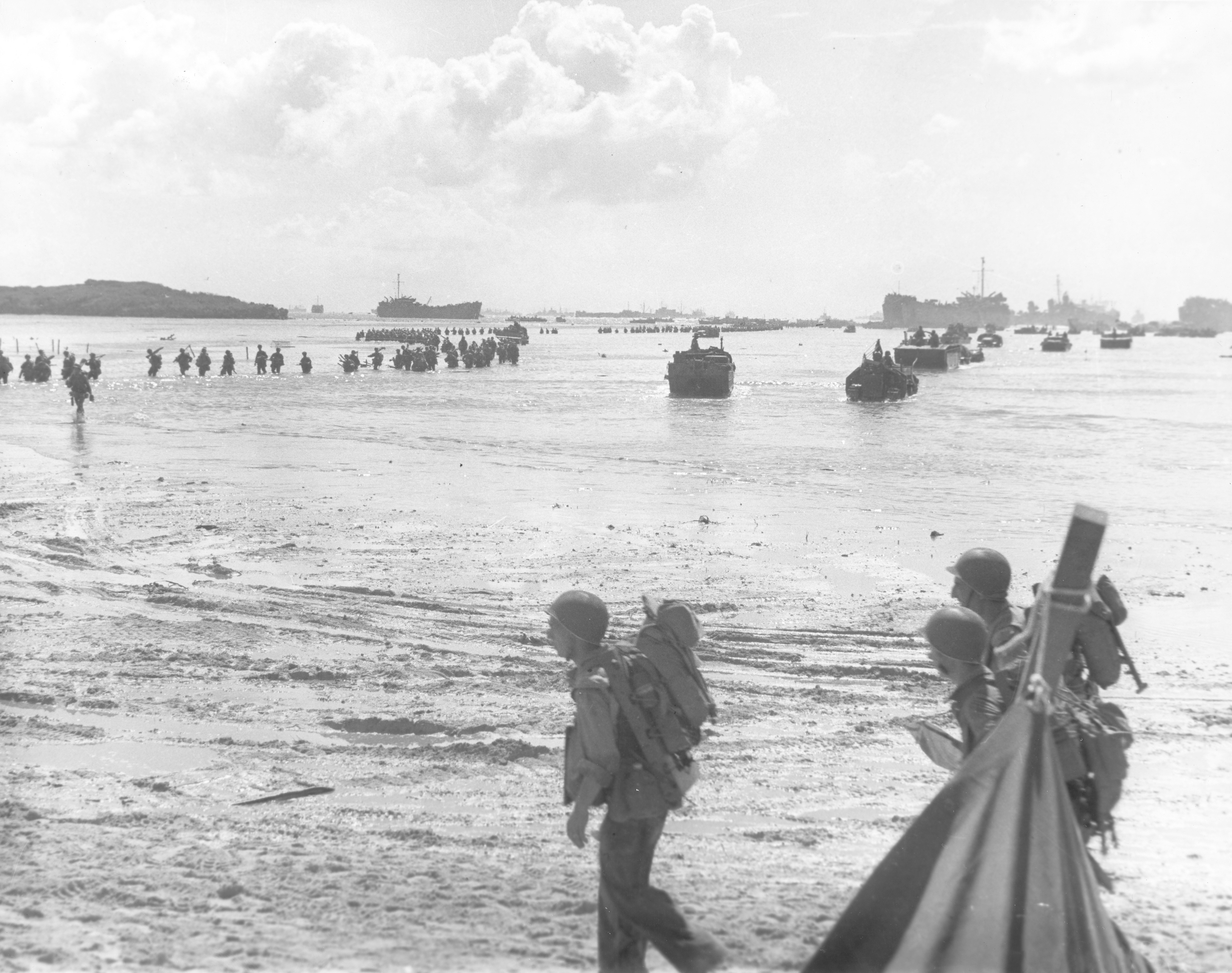 Soldiers wade through shallow waters toward the beach, with landing boats and ships in the background under a cloudy sky.