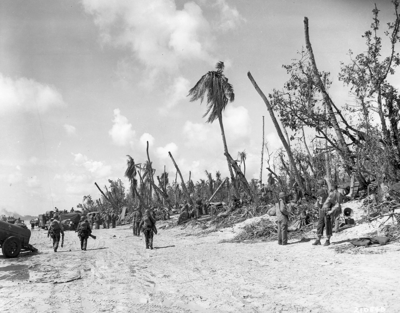Black-and-white image of soldiers on a war-torn beach, with damaged palm trees and military vehicles in the background.