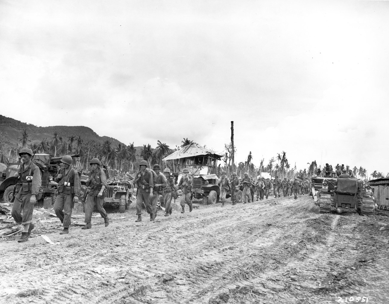 A group of soldiers in military uniforms march along a muddy road, with vehicles and a damaged building in the background amidst tropical trees.