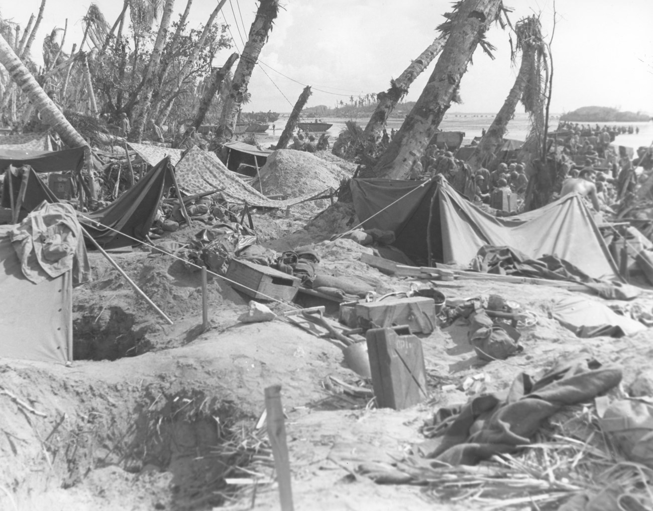 World War II battlefield scene with tents, scattered supplies, and palm trees. Soldiers gather in the background.