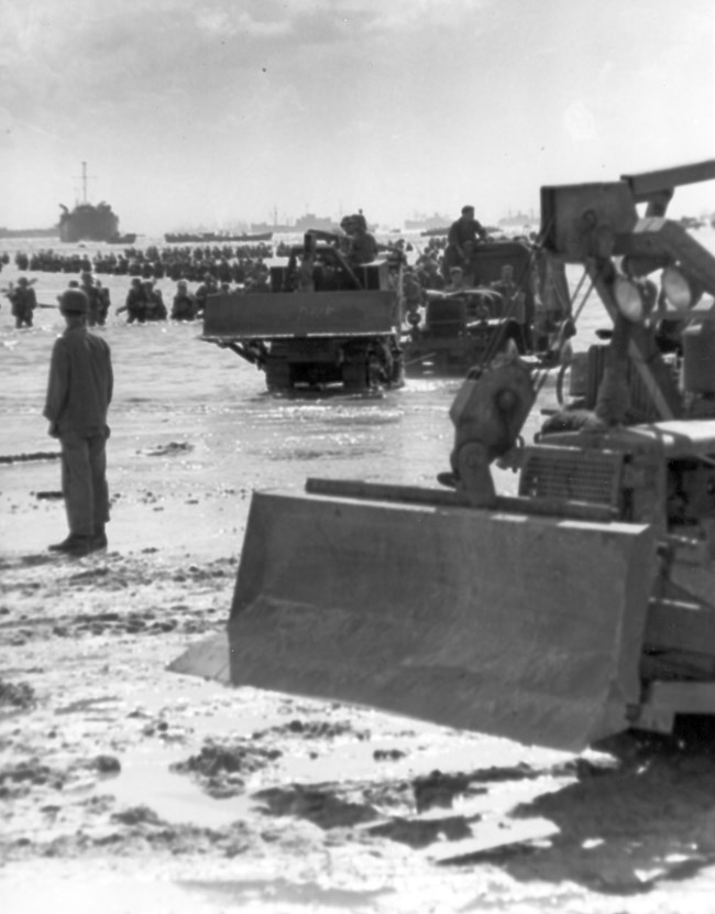 Historic WWII beach landing scene; soldiers wade ashore amid water and mud, with military trucks and a bulldozer near the foreground, conveying determination.