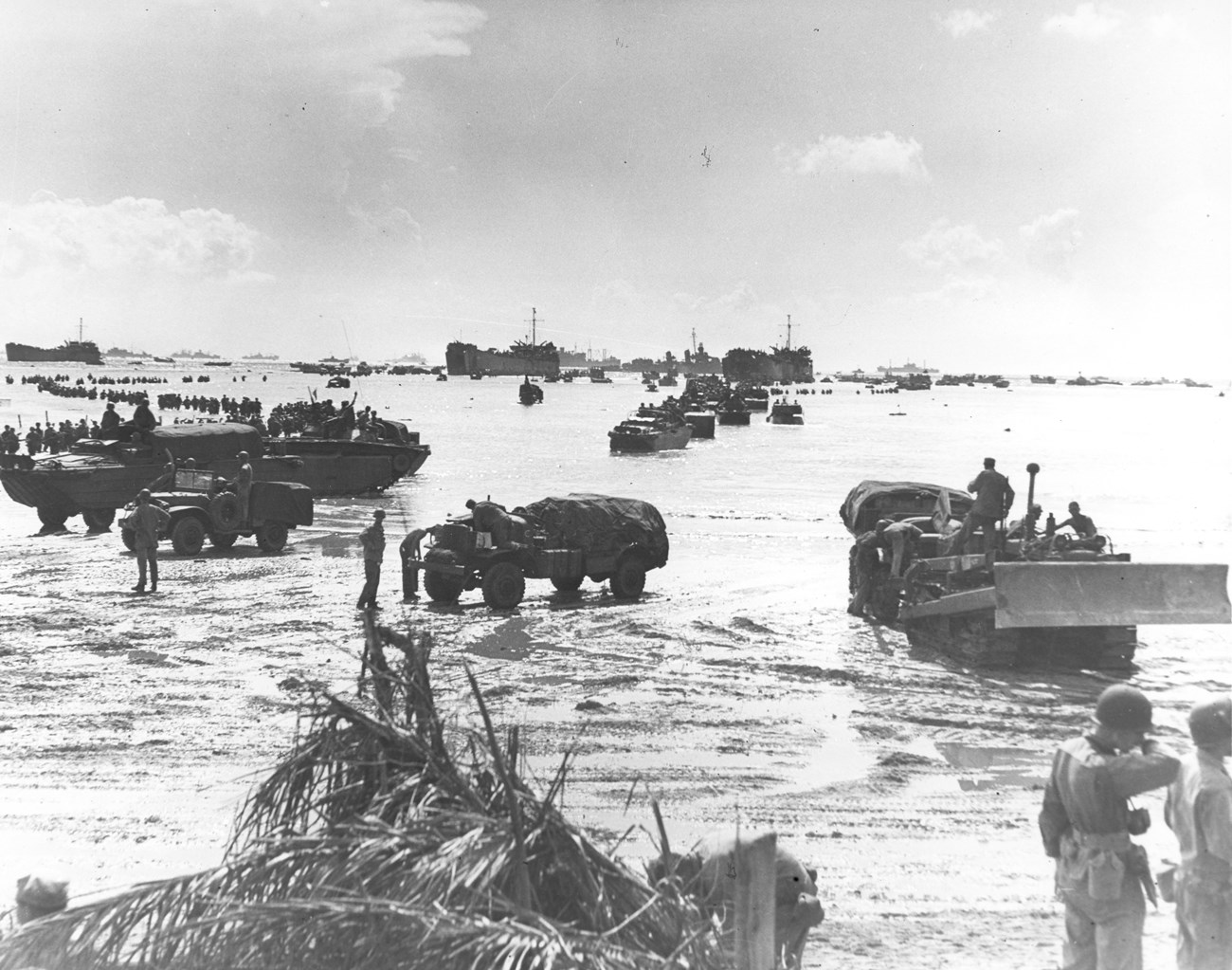 Military vehicles and troops disembark at a beach, with boats and ships in the background under a cloudy sky.