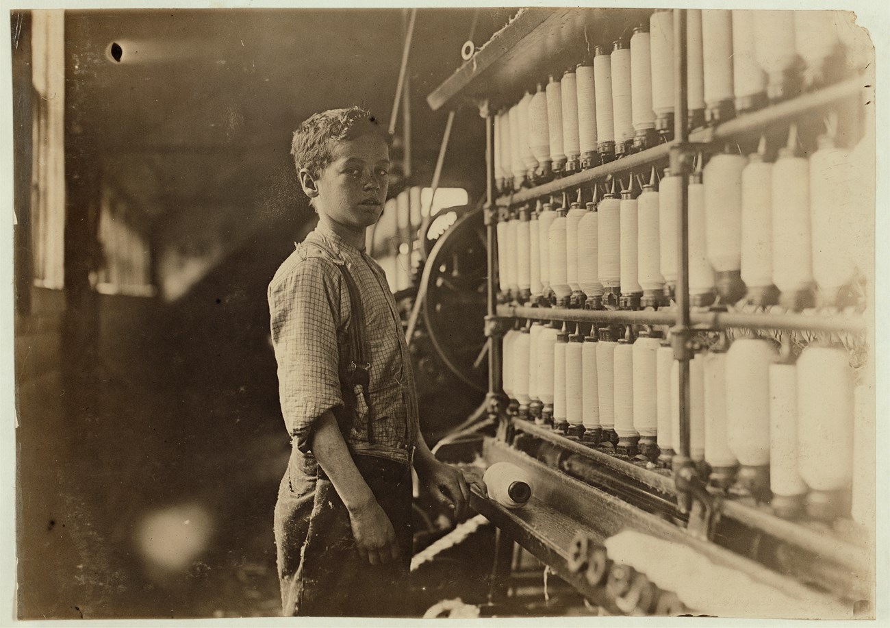 Boy standing in front of a yarn spinning machine that's taller than him, turning to face the camera.