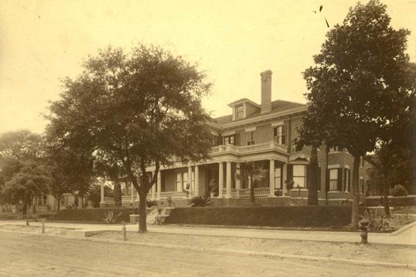 Sepia photo of a large brick house with columns and large porch surrounded by trees along the street