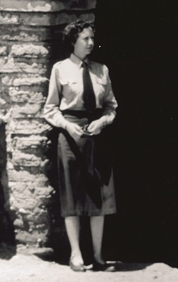 A woman stands next to a stone pillar.  She is wearing an NPS uniform.