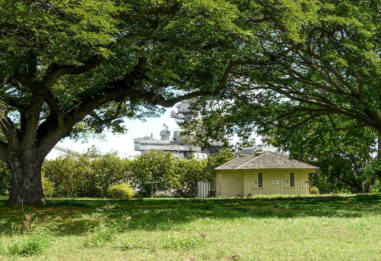 Small yellow building in grassy field dwarfed by WWII battleship in background.