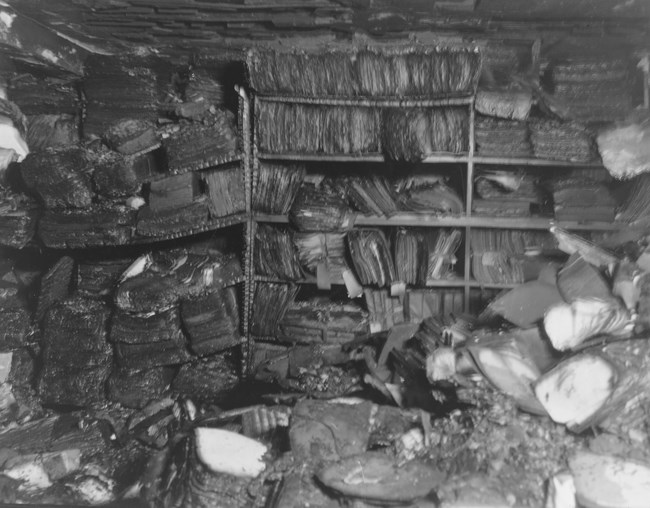 A black and white photo of paper records that have been badly burned. The records are crammed on to floor-to-ceiling shelves, with some records stacked in piles on the floor.