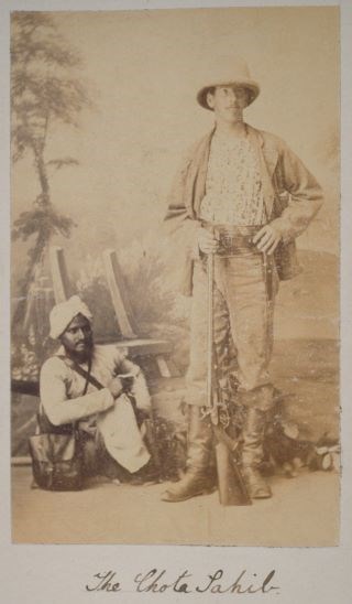 Two men pose in front of painted scene, White man standing in boots holding gun, Sikh man wearing turban sitting