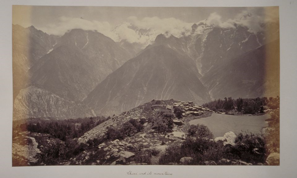Black and white photograph of small city in foothills of tall mountains with tops in snow and clouds