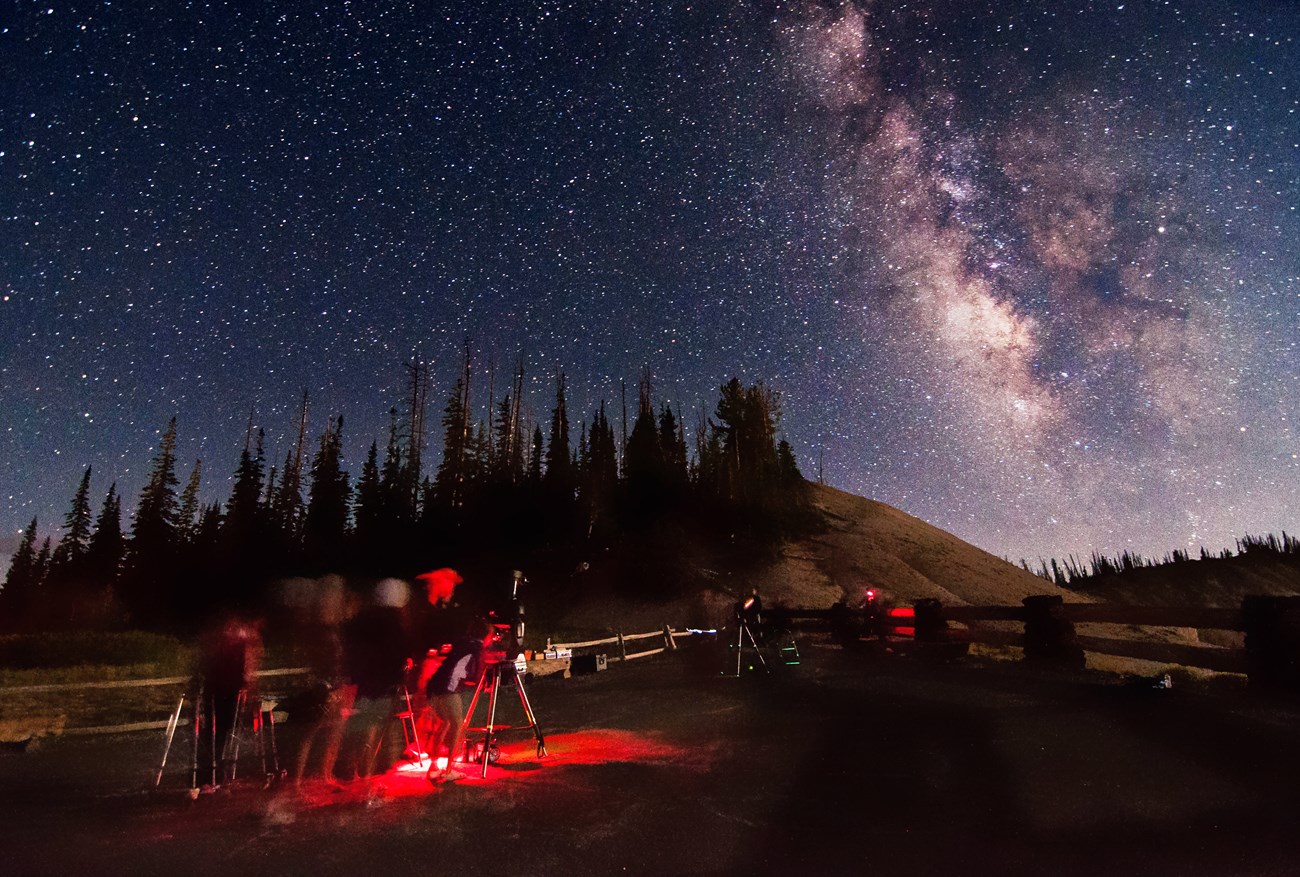 Cluster of people around telescopes, bathed in red light beneath a starry sky featuring a breathtaking view of the Milky Way galaxy.