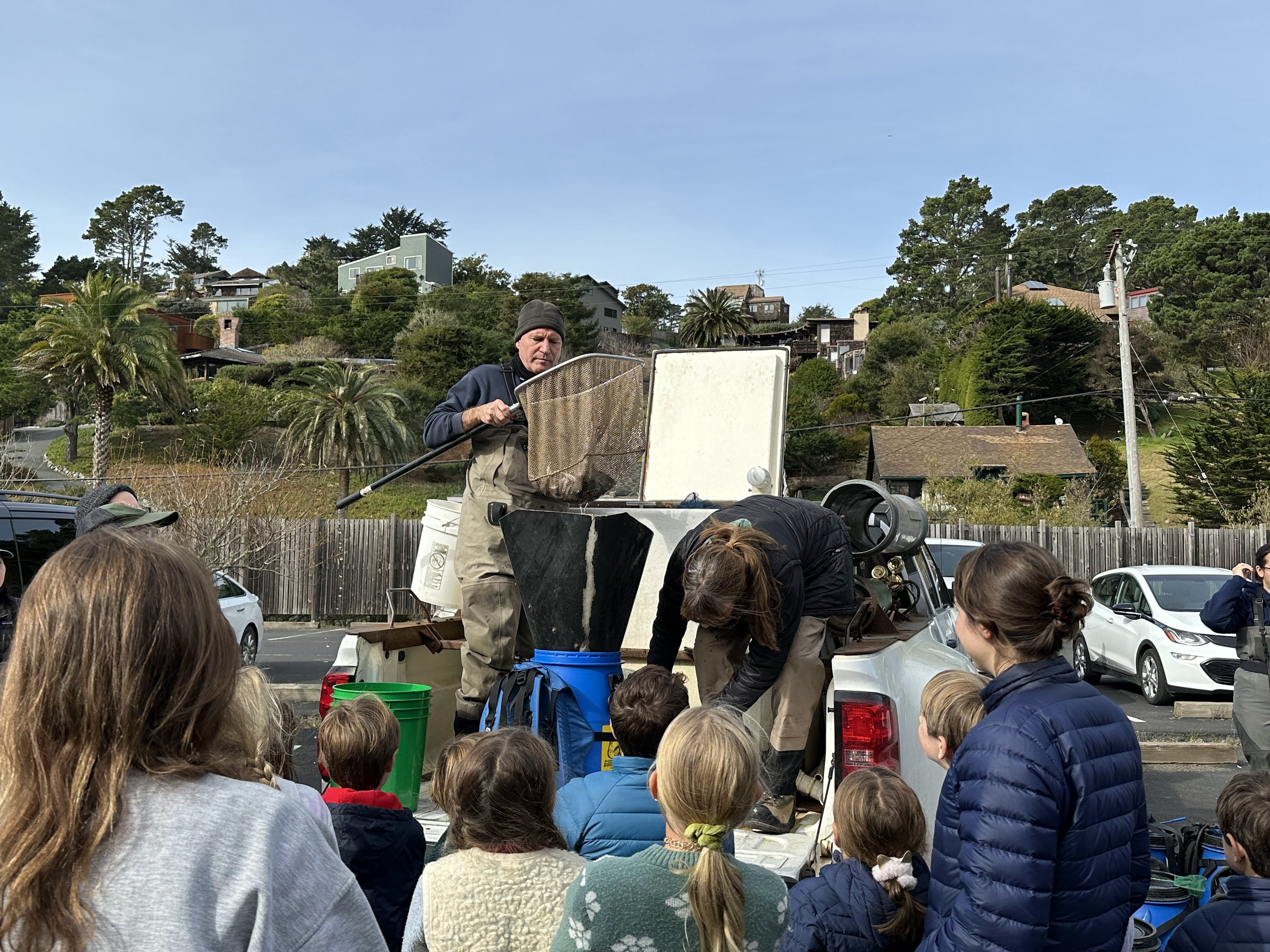 People stand on the back of a pickup truck, using a large net and funnel to transfer fish to a blue bucket-backpack in front of a crowd of children and adults.