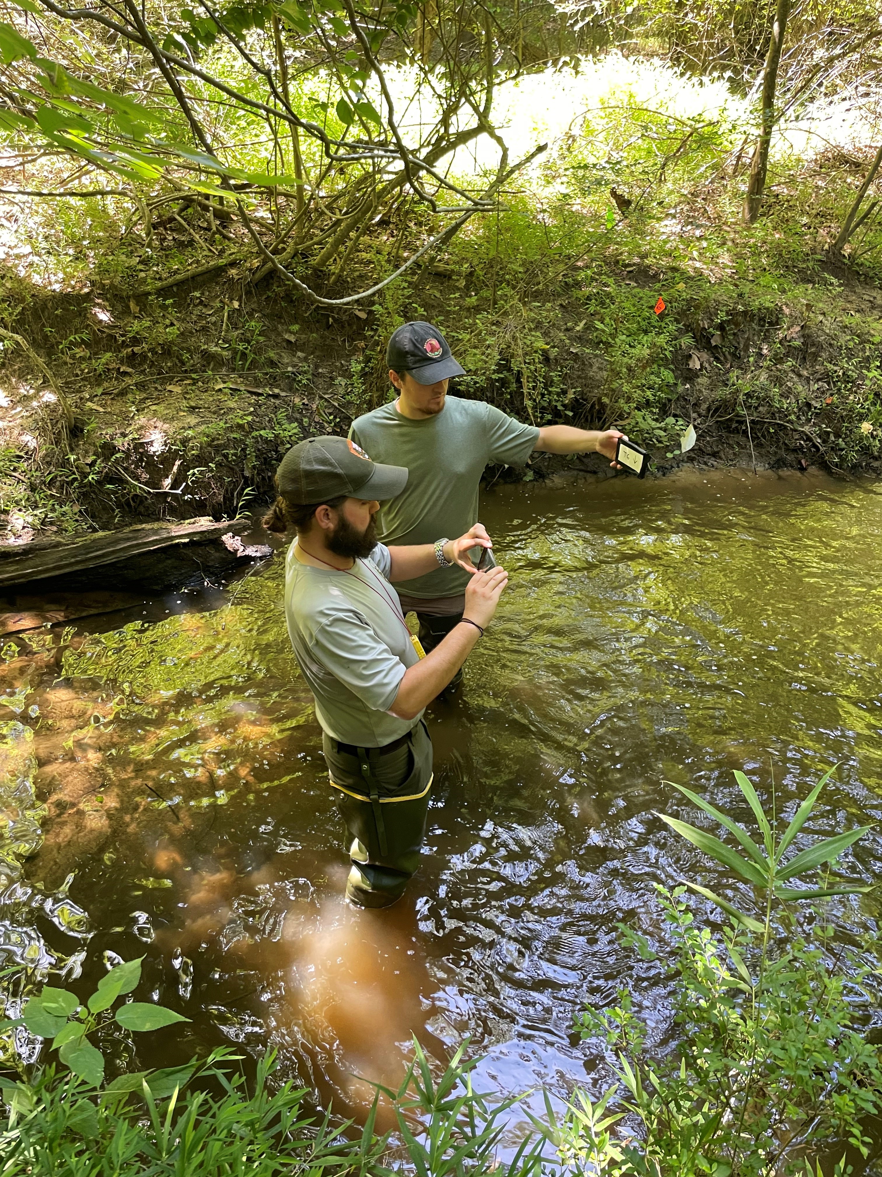 Two men standing in a creek, one holds a camera. one holds a photo card
