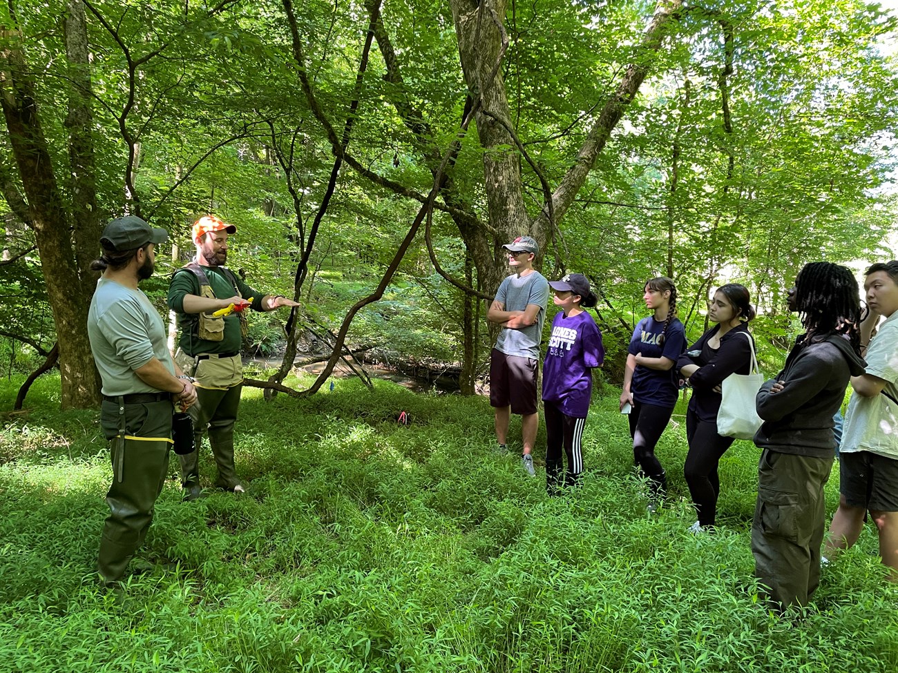 Two people, one in uniform, talk to a group of students in the forest.