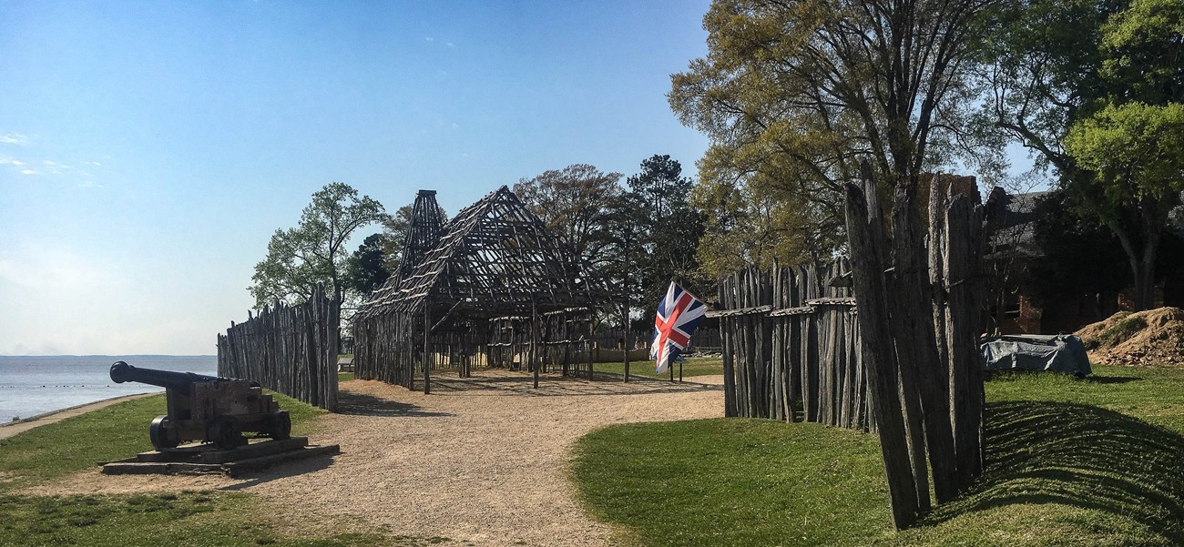 A historic building made of timber stands on the ocean shorelinr behind stockade fencing and a large cannon and British flag.