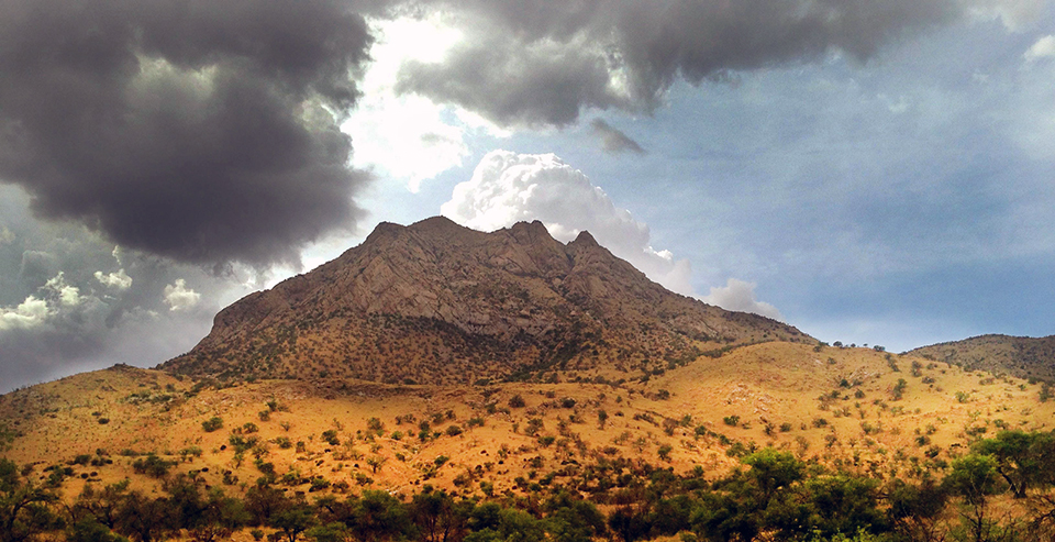 Sunlight peeking through dark storm clouds over a barren mountain surrounded by desert hills covered in dry grasses and green shrubs.
